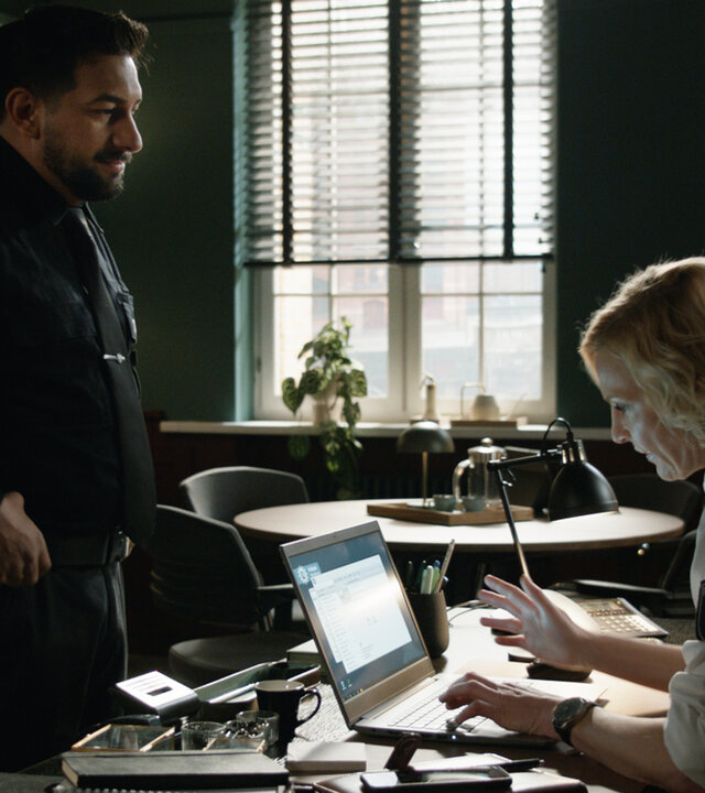 "Notruf Hafenkante - Vertrauensbruch": Die Polizistin Melanie Hansen (Sanna Englund) sitzt in ihrem Büro an ihrem Schreibtisch. Sie tippt etwas in einen Laptop, der geöffnet vor ihr steht. Vor ihrem Schreibtisch steht ihr Kollege Malik (Zejhun Demirov). Er grinst sie an.