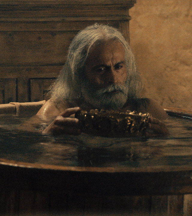 "Die glorreichen 10 - Die krassesten Kräfte der Geschichte": Spielszene - Archimedes sitzt mit überlegendem Blick in einer randvoll gefüllten Badewanne. Er schaut auf eine Goldene Krone, die er auf der Wasseroberfläche hält.