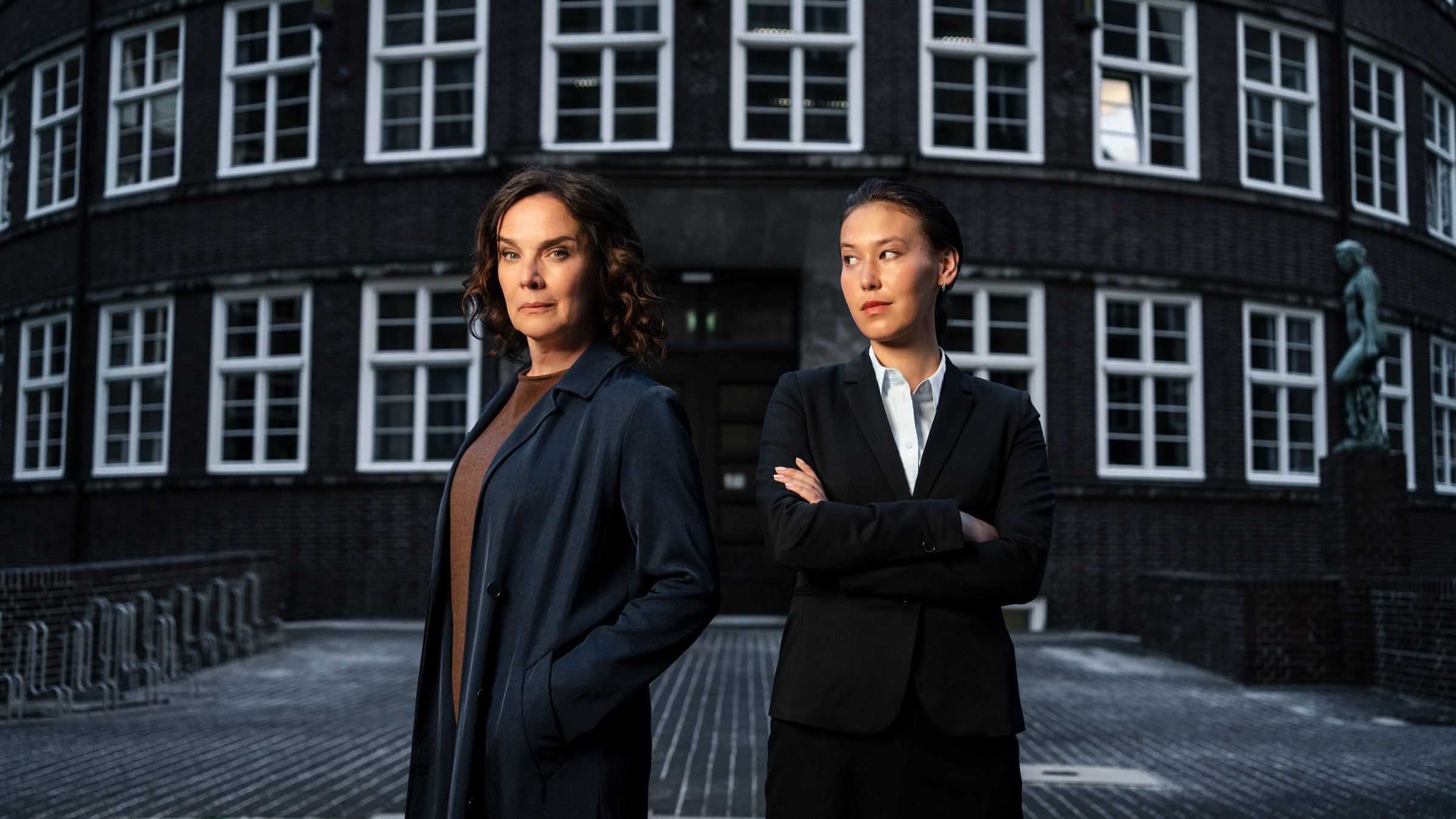 "Conti - Meine zwei Gesichter": Doppelporträt von Désirée Nosbusch und Malaya Stern Takeda in ihrer jeweiligen Rolle als Rechtsanwältin Anna Conti und Staatsanwältin Henry Mahn. Die beiden Schauspielerinnen stehen Schulter an Schulter vor einem Backsteingebäude mit weißen Fenstern.