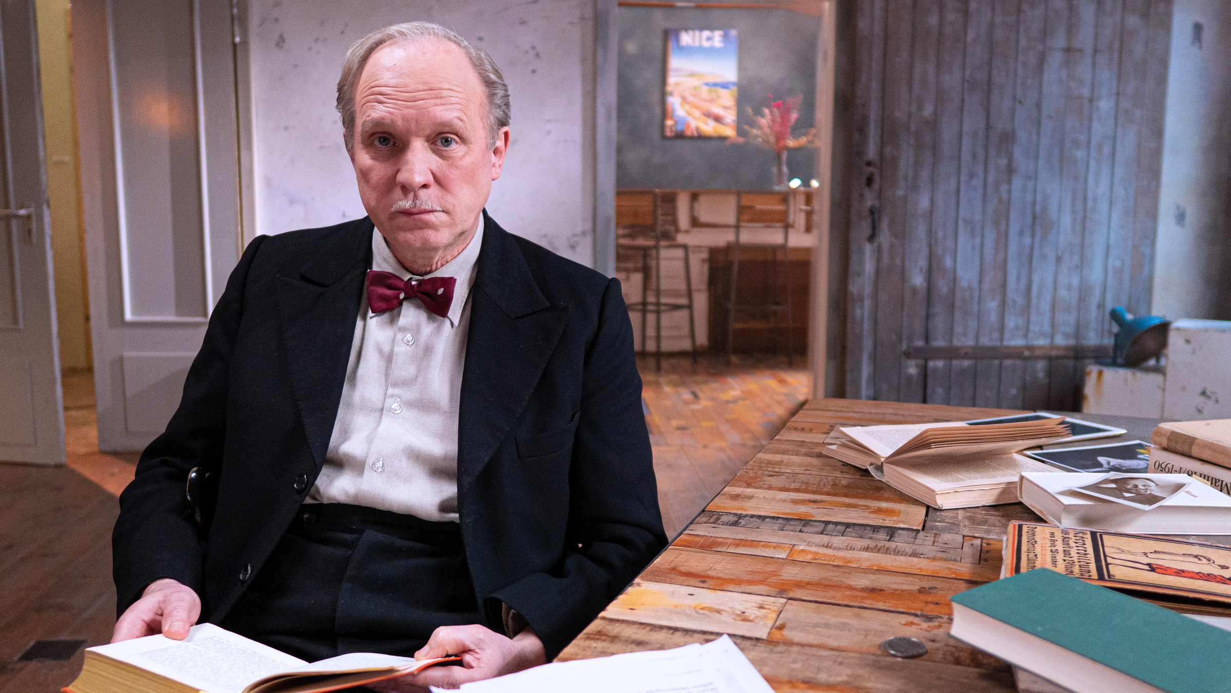 „Heinrich Mann, der unbekannte Rebell:“ Der Schauspieler Ulrich Tukur sitzt an einem Schreibtisch, um ihn herum viele Bücher. Ein Buch hält er in die Hand, er schaut in die Kamera.