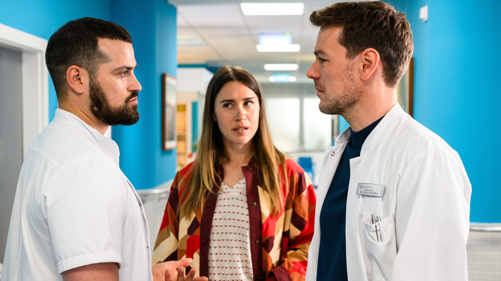 "Bettys Diagnose - Echte Vorurteile": Tarik (Nicolas Fethi Türksever) und Julius (Constantin Lücke) stehen sich im Krankenhausflur gegenüber und schauen sich an. Betty (Henrike Hahn) steht zwischen den beiden und schaut dabei Tarik an.