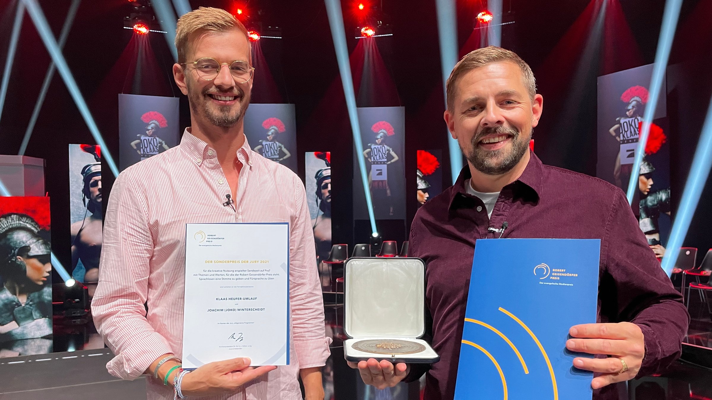 "Der Robert Geisendörfer Preis 2021": Joko Winterscheidt (links) und Klaas Heufer-Umlauf (rechts) halten jeder in der Hand eine Urkunde und in der Mitte wird von ihnen der Robert-Geisendörfer-Preis, eine große Medaille, in einer Schatulle präsentiert.