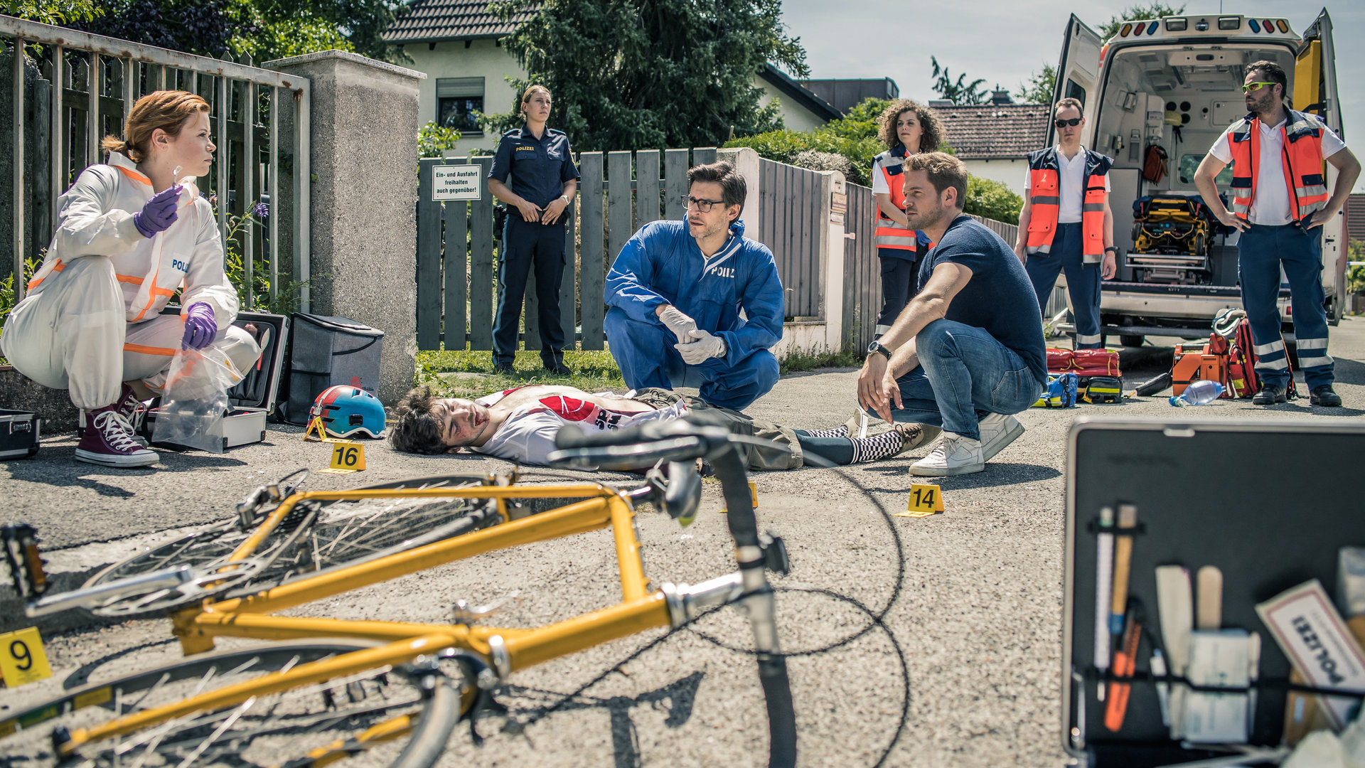 "SOKO München - Die letzte Lieferung": Auf der Straße in einem Wohngebiet liegt eine männliche Leiche, etwas entfernt das Fahrrad, mit dem der Unfall passiert ist. Dr. Weissenböck (Florian Odendahl) und Dominik Morgenstern (Joscha Kiefer) sitzen in der Hocke neben der Leiche und schauen zu Billie Curio (Sina Reiß), die auf dem Bürgersteig Beweismaterial mit einer Pinzette hochhält. Im Hintergrund stehen eine Polizistin und ein Sanitäter-Team (Darsteller unbekannt).