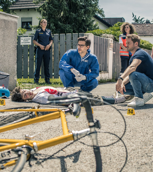 "SOKO München - Die letzte Lieferung": Auf der Straße in einem Wohngebiet liegt eine männliche Leiche, etwas entfernt das Fahrrad, mit dem der Unfall passiert ist. Dr. Weissenböck (Florian Odendahl) und Dominik Morgenstern (Joscha Kiefer) sitzen in der Hocke neben der Leiche und schauen zu Billie Curio (Sina Reiß), die auf dem Bürgersteig Beweismaterial mit einer Pinzette hochhält. Im Hintergrund stehen eine Polizistin und ein Sanitäter-Team (Darsteller unbekannt).
