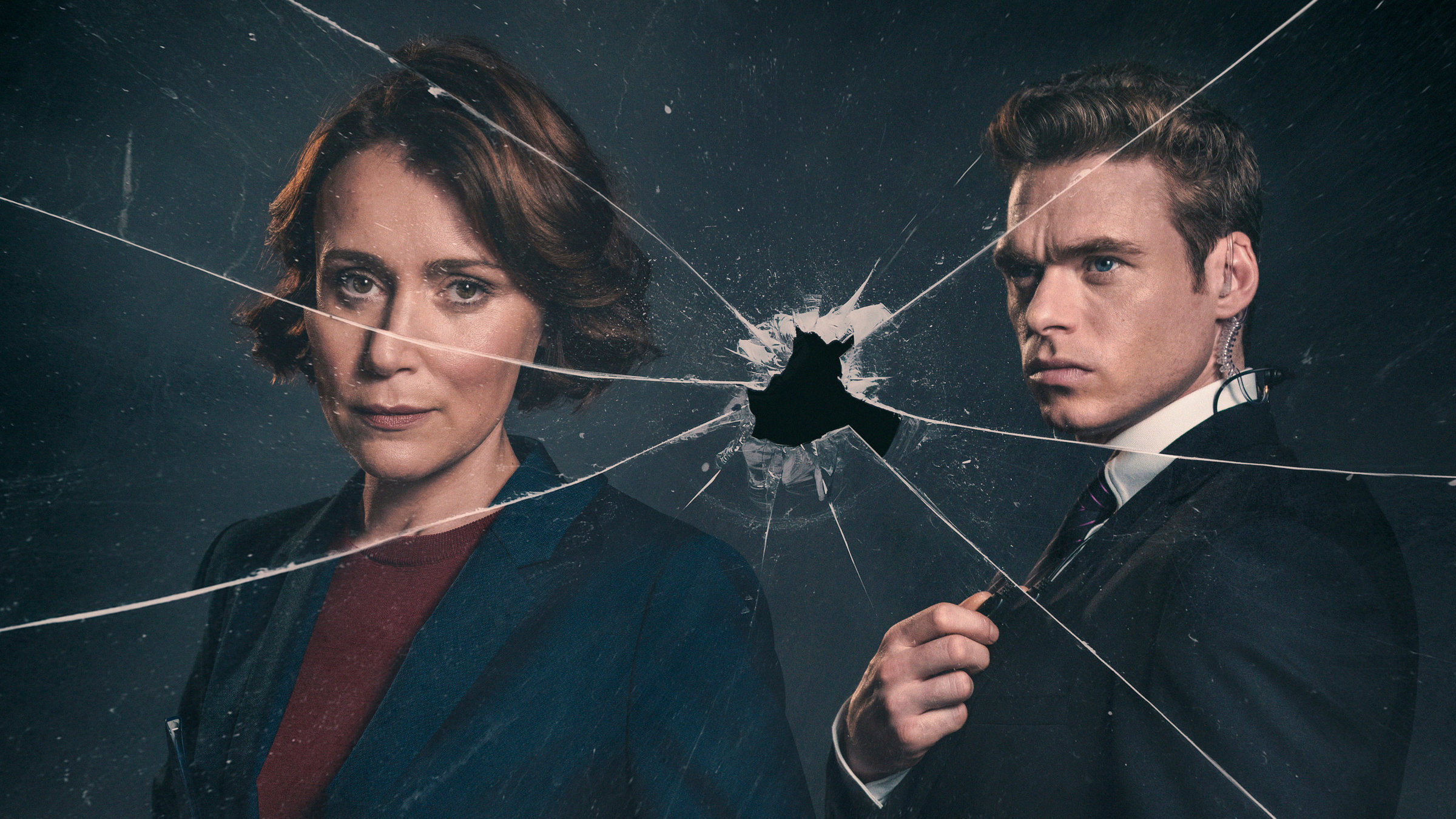 "Bodyguard": Julia (Keeley Hawes) und David (Richard Madden) stehen hinter einer gesprungenen Glasscheibe, die ein Einschussloch in der Mitte hat. David hat sein Funkgerät in der Hand.
