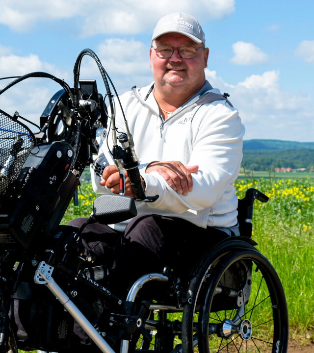 "einfach Mensch - Christian Nachtwey: Von Vielen unterschätzt (3)": Christian Nachtwey sitzt auf dem Handbike an einem Wiesenrand bei Sonnenschein. Er blickt lächelnd in die Kamera.