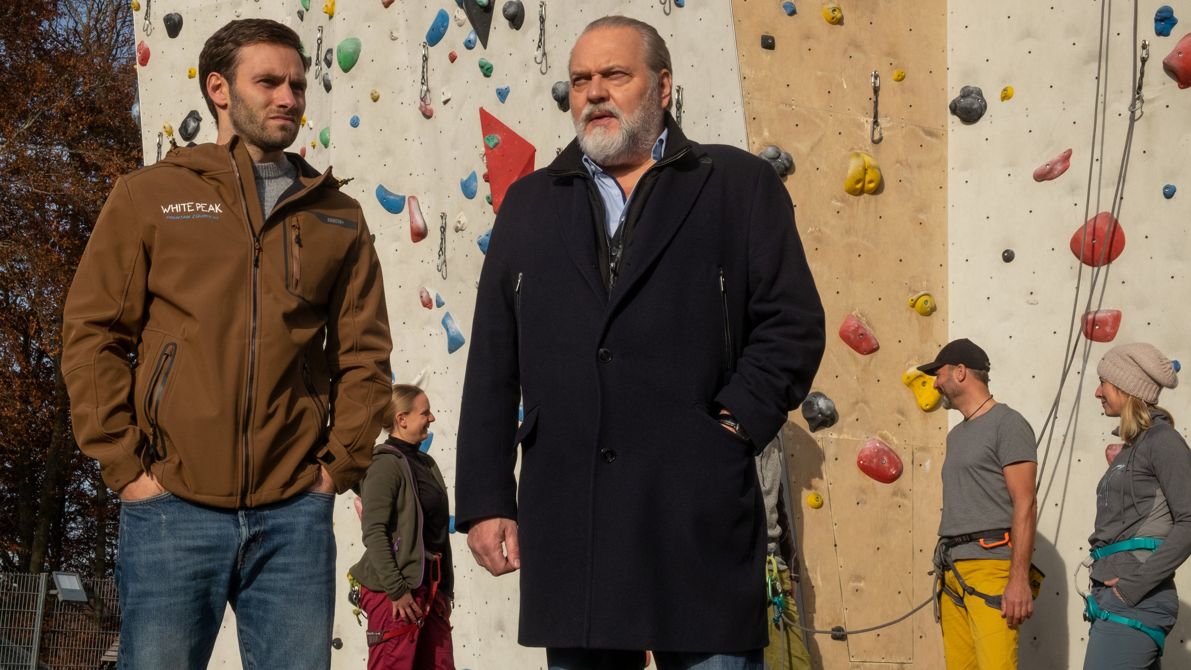 "SOKO Leipzig - Freier Fall": Eric Du Bois (Jonathan Müller) steht mit Hauptkommissar Richard Voss (Jan-Gregor Kremp) im Freien vor einer Kletterwand. Im Hintergrund stehen einige Personen, die sich zum Klettern getroffen haben.