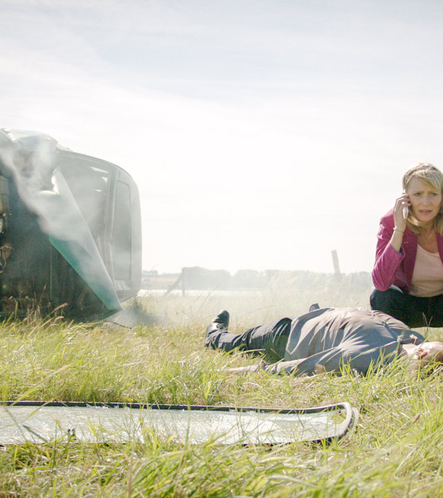 „SOKO Wismar – Aus der Kurve“: Roswitha Prinzler (Silke Matthias) hockt auf einer Wiese und telefoniert. Vor ihr liegt der reglose Jan Lehmann (Christopher Fliether). Etwas daneben liegt ein demolierter Wagen auf die Fahrerseite gekippt, im Hintergrund ist eine Landstraße.