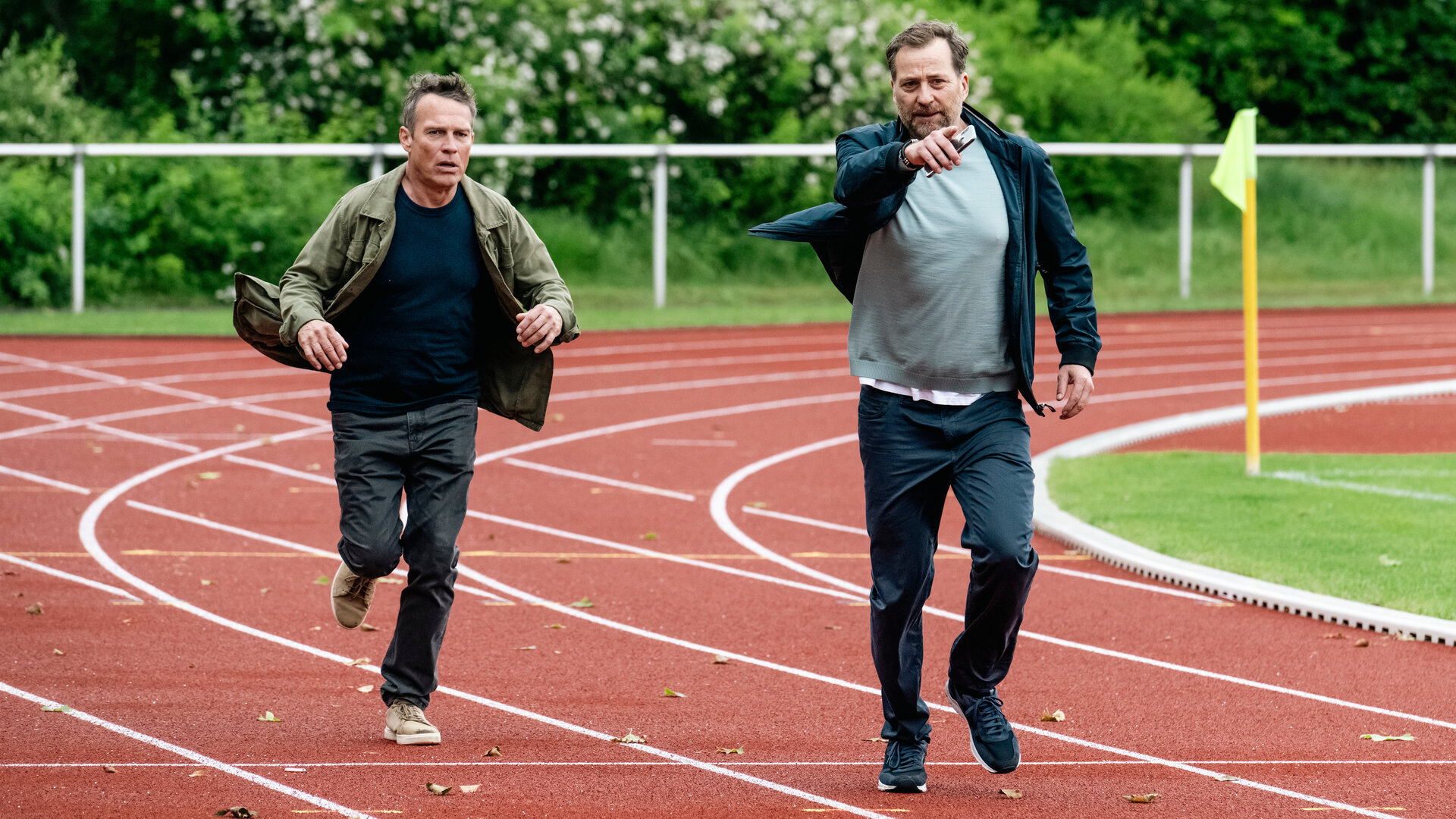 "SOKO Köln - Ins Abseits gelaufen": Matti Wagner (Pierre Besson) rennt neben Thomas Frysell (Christian Erdmann) über die Laufbahn auf einem Sportplatz. Thomas Frysell deutet mit der rechten Hand, in der er sein Handy hält, nach vorne. Matti sieht mit ernstem Blick zu der Stelle.