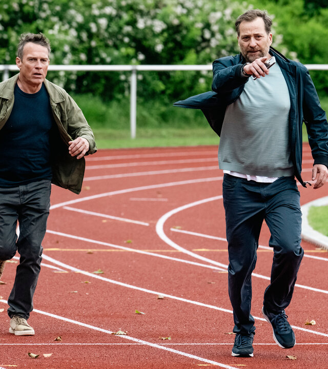 "SOKO Köln - Ins Abseits gelaufen": Matti Wagner (Pierre Besson) rennt neben Thomas Frysell (Christian Erdmann) über die Laufbahn auf einem Sportplatz. Thomas Frysell deutet mit der rechten Hand, in der er sein Handy hält, nach vorne. Matti sieht mit ernstem Blick zu der Stelle.