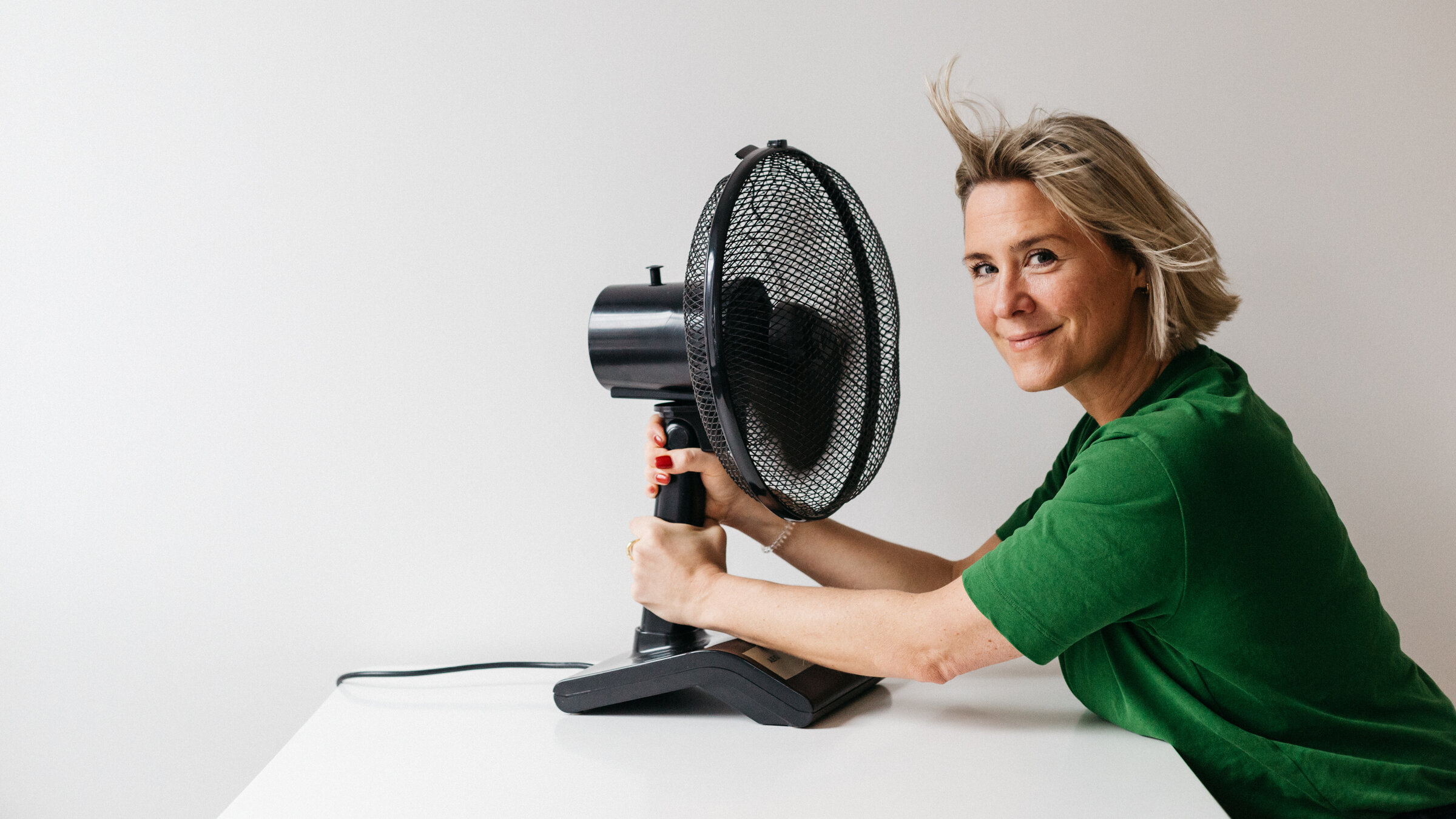 "Wechseljahre: heißkalt erwischt": Line sitzt vor einer weißen Wand an einem weißen Tisch. Sie hält einen schwarzen Tischventilator fest und schaut lächelnd in die Kamera.