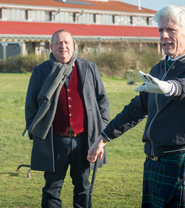 "SOKO Wismar – Der Golfer mit dem Schottenrock": Im Hintergrund ist das große Gebäude des Golfplatzes zu sehen. Auf dem Golfplatz selbst steht Jan Reuter (Udo Kroschwald), der mit Golfer Ian MacPherson (Philippe Brenninkmeyer) spricht. Der Golfer trägt Golferhandschuhe und einen Quilt und hält mit der rechten Hand einen Golfschläger fest.