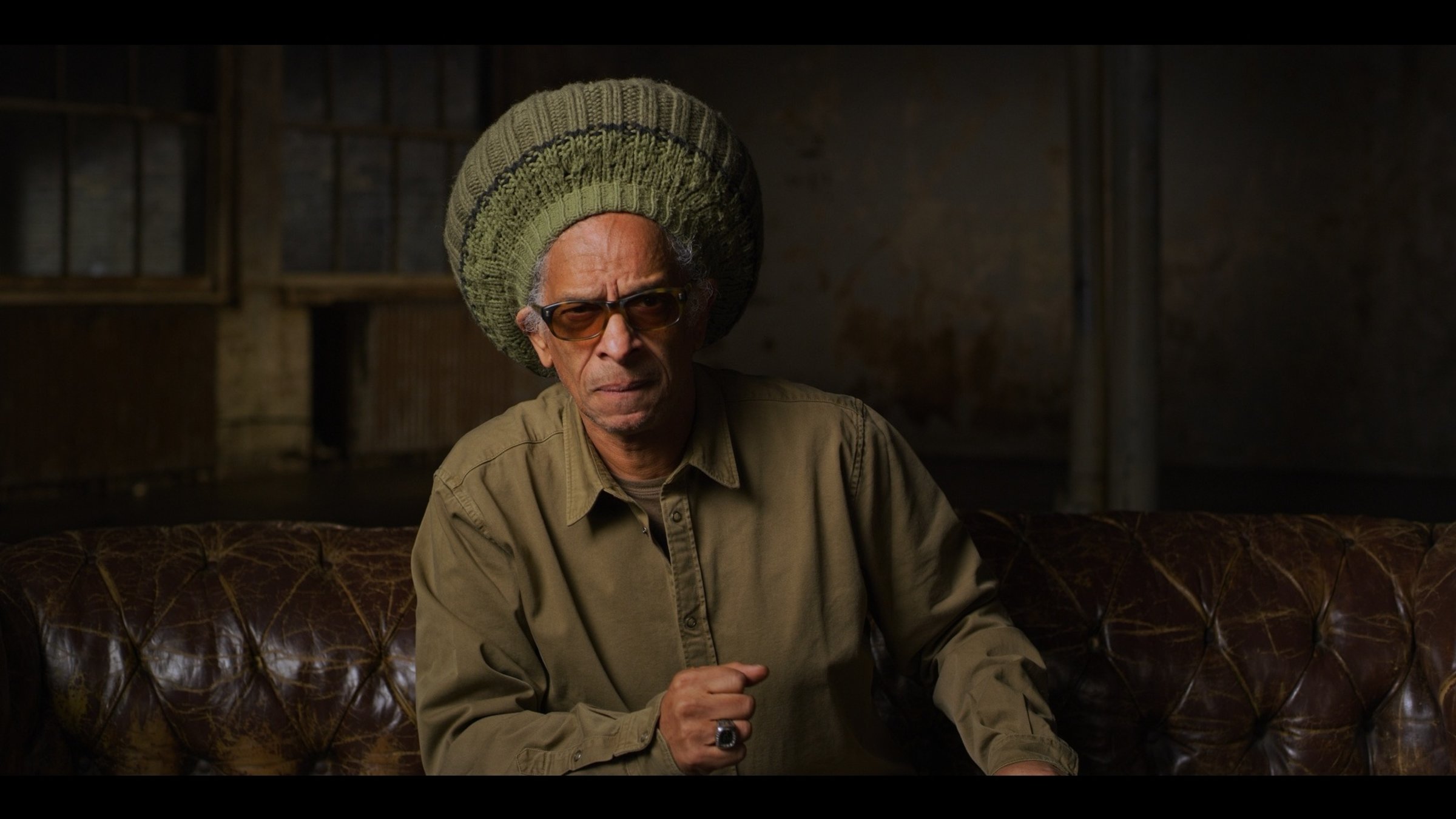 "The True Story of Punk - Die Welle": Don Letts sitzt auf einem braunen Ledersofa und schaut in die Kamera. Er trägt eine große olivfarbene Mütze und eine Sonnenbrille.