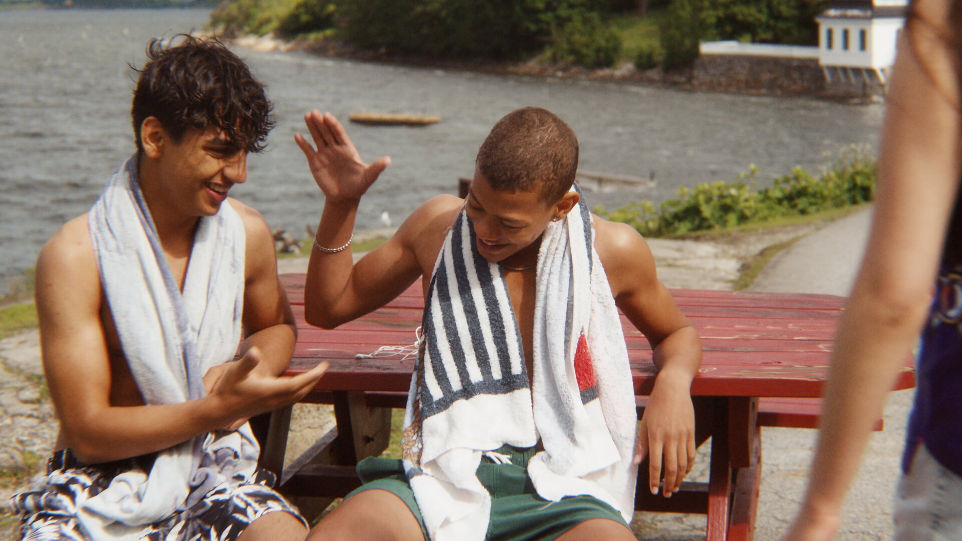 "After Benjamin: Holmlia For Life": Benjamin (Emil Sesay Stenseth) und Elias (Sam Ashraf) sitzen in Badekleidung am Osloer Meeresufer vor einem roten Holztisch auf einer Bank und klatschen sich ab. Sie wirken entspannt und gut gelaunt.
