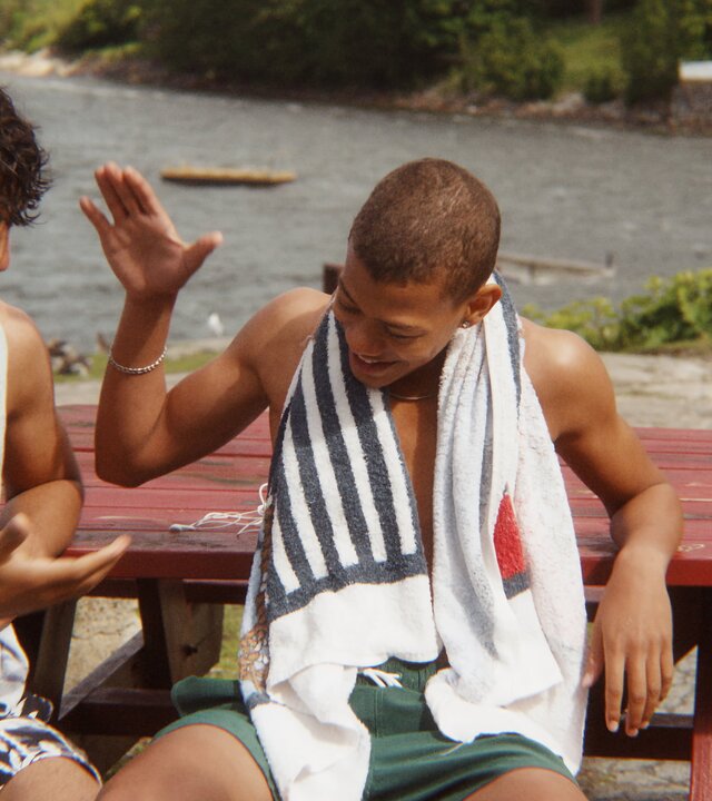 "After Benjamin: Holmlia For Life": Benjamin (Emil Sesay Stenseth) und Elias (Sam Ashraf) sitzen in Badekleidung am Osloer Meeresufer vor einem roten Holztisch auf einer Bank und klatschen sich ab. Sie wirken entspannt und gut gelaunt.