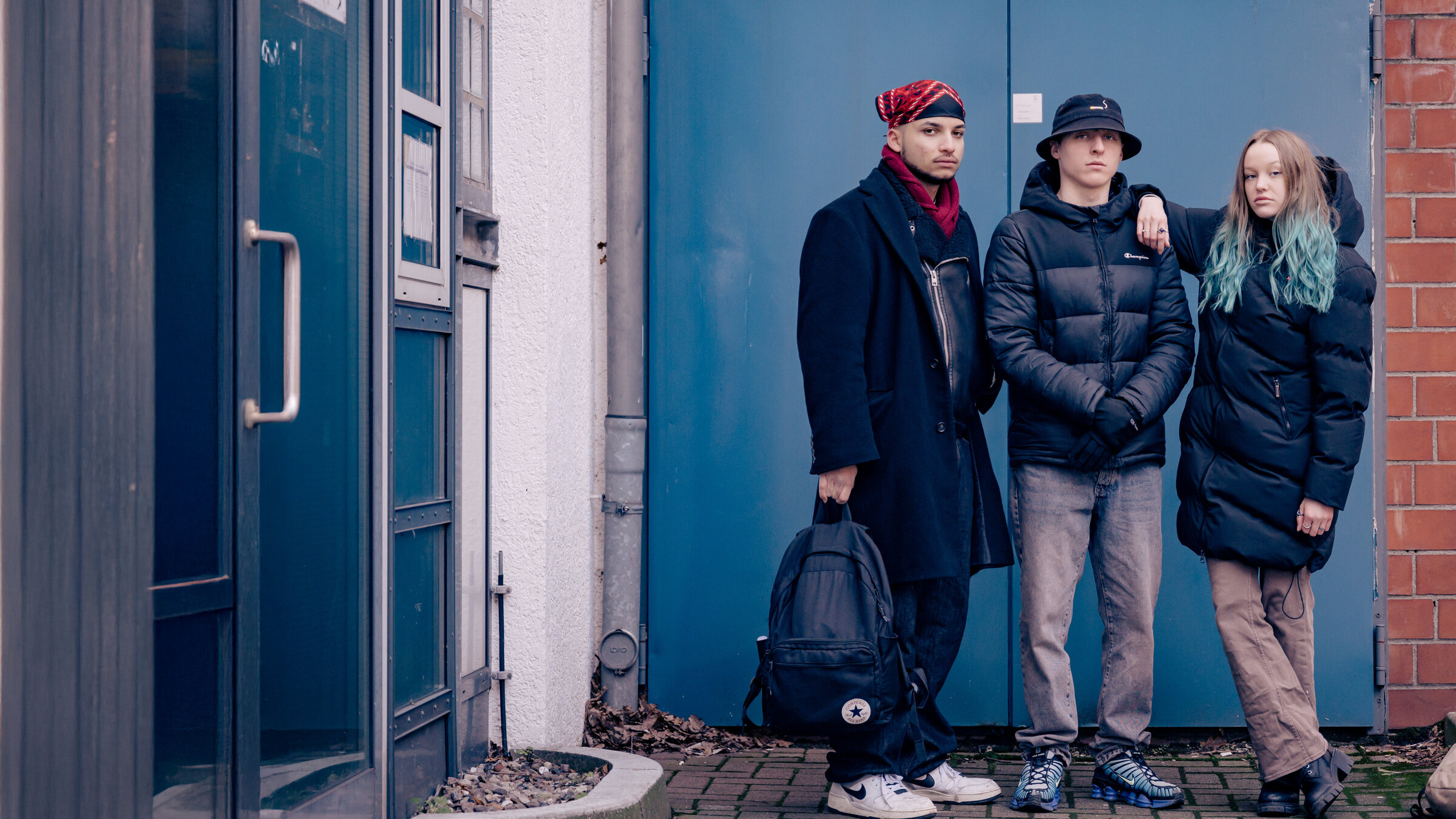"Letzte Chance für Schulabbrecher": Merlin, Angelo und Liska stehen vor einer blauen Stahltür. Alle schauen in die Kamera.