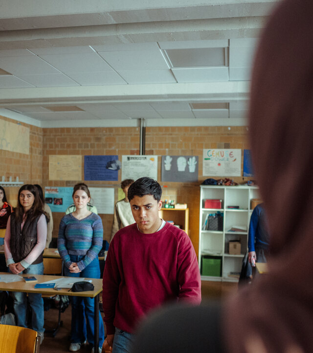 "Uncivilized - Nine Eleven": Ahmad (Rasmi Mohammed Nasrallah) verlässt wütend den Klassenraum, während seine Mitschülerinnen und Mitschüler mit gefalteten Händen an Ihren Plätzen stehen.