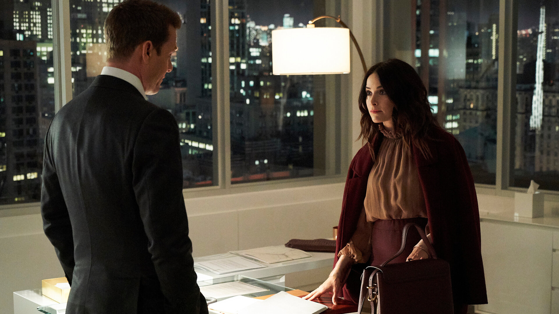 "Suits - Vom gleichen Schlag": Dana (Abigail Spencer) steht am Schreibtisch und schaut den vor dem Schreibtisch stehenden Harvey (Gabriel Macht) an, dessen Rücken der Kamera zugewandt ist. Sie trägt ein elegantes, dunkelrotes Ensemble mit mokkafarbener Bluse. Vor ihr auf dem Tisch steht eine farblich abgestimmte große Handtasche.