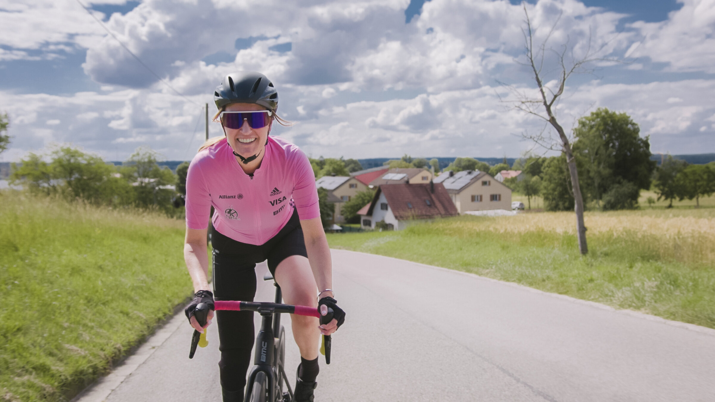 "einfach Mensch - Denise Schindler: Paralympics 2024 (1)": Denise Schindler fährt auf ihrem Rennrad eine Straße hoch. Sie lacht in die Kamera. Im Hintergrund liegen Wiesen, dahinter stehen Häuser unter einem bewölkten Himmel.