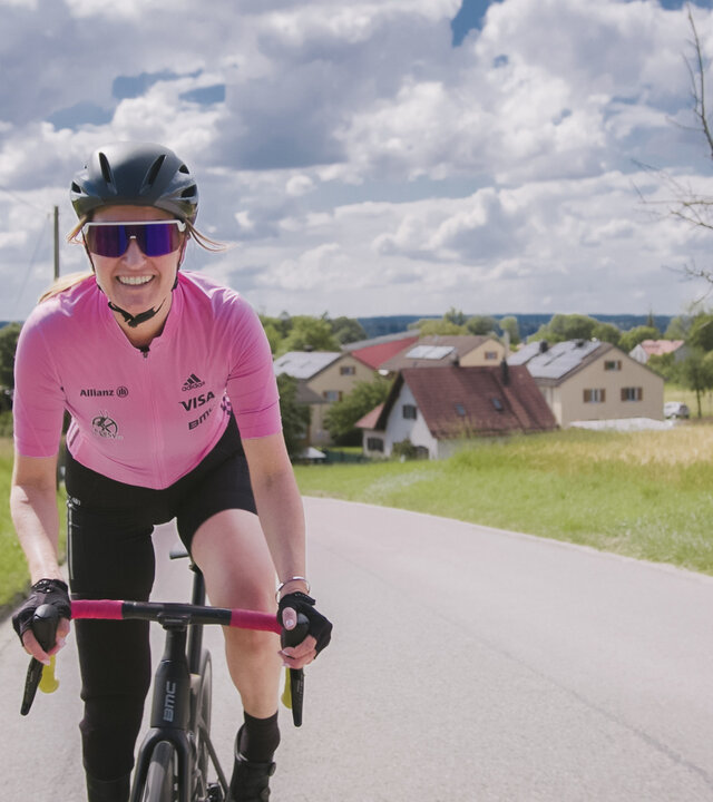 "einfach Mensch - Denise Schindler: Paralympics 2024 (1)": Denise Schindler fährt auf ihrem Rennrad eine Straße hoch. Sie lacht in die Kamera. Im Hintergrund liegen Wiesen, dahinter stehen Häuser unter einem bewölkten Himmel.