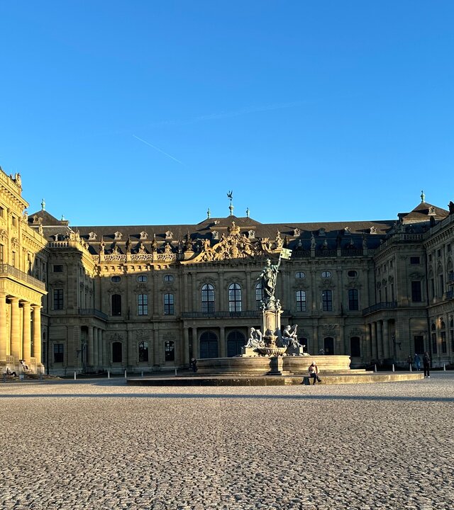"Abschlussgottesdienst zum Katholikentag - Hab Mut, steh auf ...und geh!": Unter blauem Himmel liegt der gepflasterte Residenzplatz, dahinter dahinter liegt die Würzburger Residenz, ein Barockschloss.