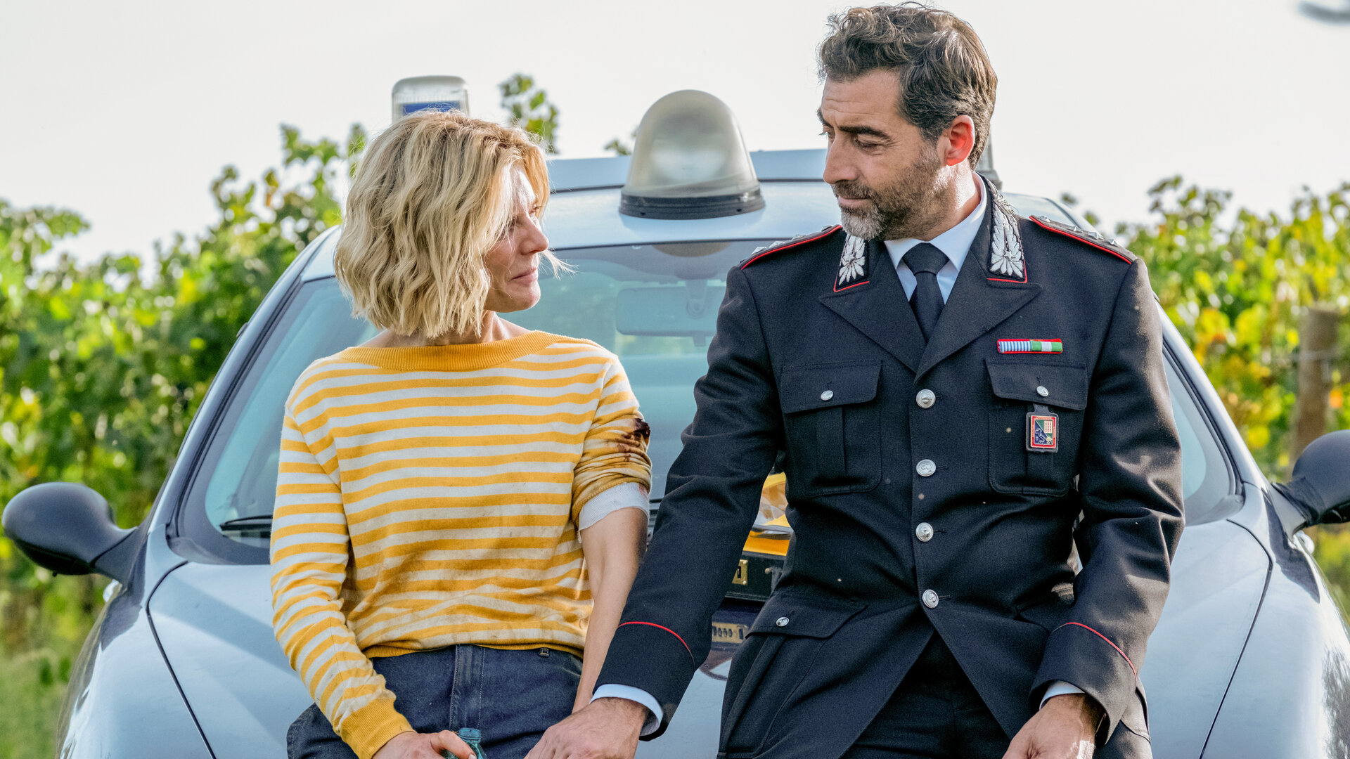 "Signora Volpe - Familiensache": Sylvia (Emilia Fox) und Giovanni (Giovanni Cirfiera) sitzen nebeneinander auf der Motorhaube eines Polizeiautos. Er trägt seine Uniform als Capitano. Die beiden halten Händchen.