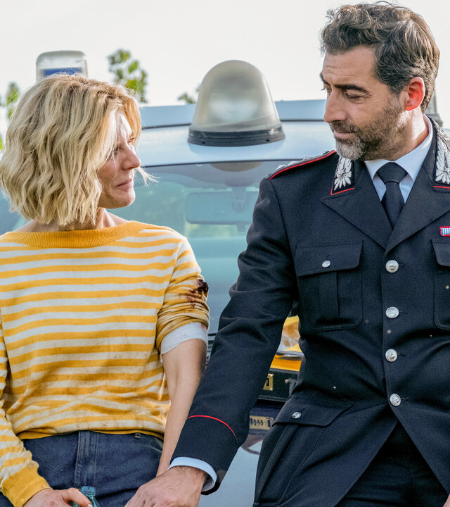 "Signora Volpe - Familiensache": Sylvia (Emilia Fox) und Giovanni (Giovanni Cirfiera) sitzen nebeneinander auf der Motorhaube eines Polizeiautos. Er trägt seine Uniform als Capitano. Die beiden halten Händchen.