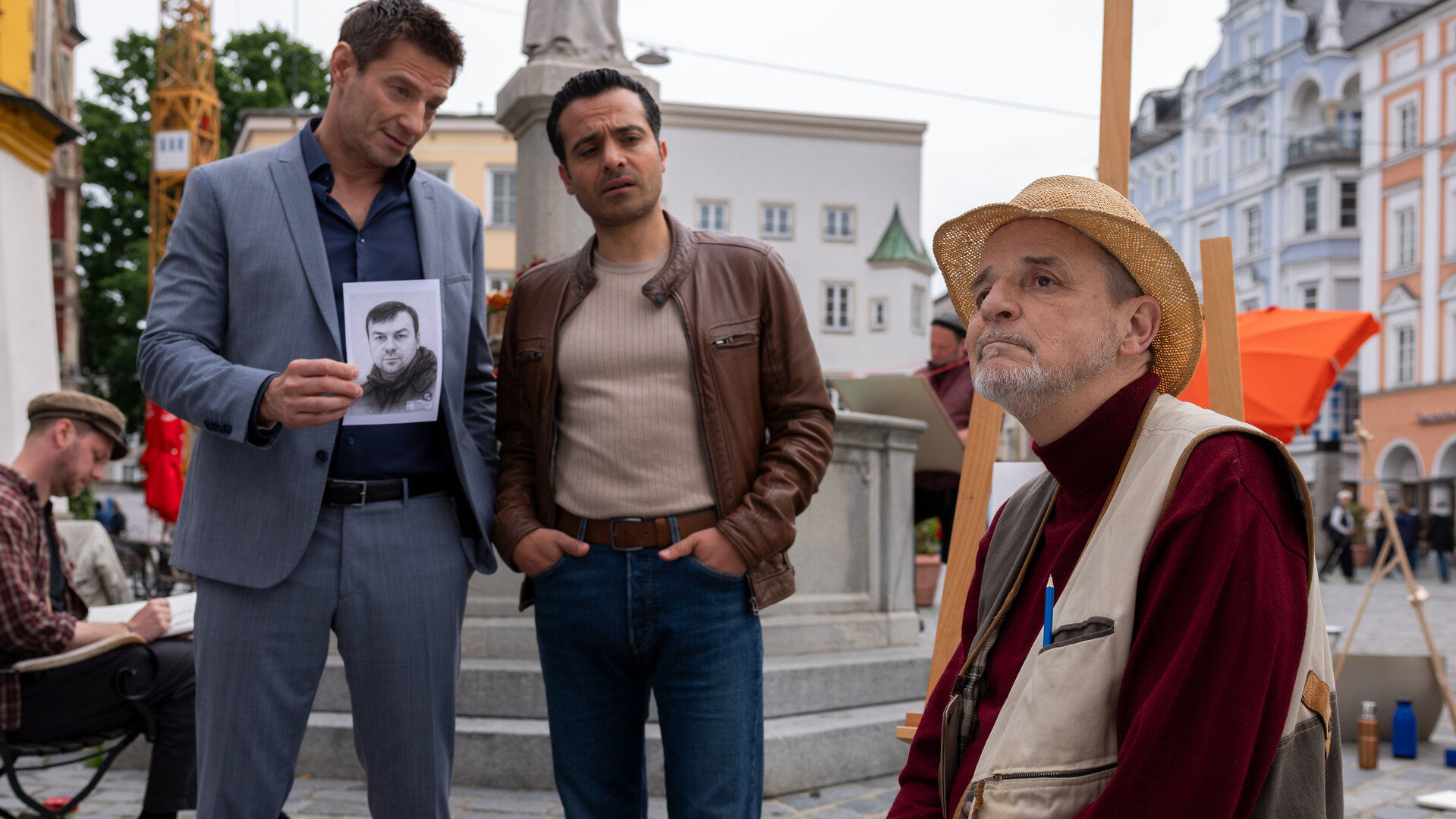 "Die Rosenheim-Cops - Die Phantomzeichnung": Die Kommissare Hansen (Igor  Jeftić) und Kaya (Baran Hêvî) stehen nebeneinander in der Innenstadt von Rosenheim vor einem Brunnen und schauen Oswald Greimer (Christian Bruse) an, der vor ihnen auf einem Stuhl sitzt. Greimer trägt einen Strohhut und blickt abwesend in die Ferne. Im Hintergrund sind weitere Personen auf Stühlen zu sehen.
