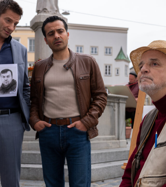 "Die Rosenheim-Cops - Die Phantomzeichnung": Die Kommissare Hansen (Igor  Jeftić) und Kaya (Baran Hêvî) stehen nebeneinander in der Innenstadt von Rosenheim vor einem Brunnen und schauen Oswald Greimer (Christian Bruse) an, der vor ihnen auf einem Stuhl sitzt. Greimer trägt einen Strohhut und blickt abwesend in die Ferne. Im Hintergrund sind weitere Personen auf Stühlen zu sehen.