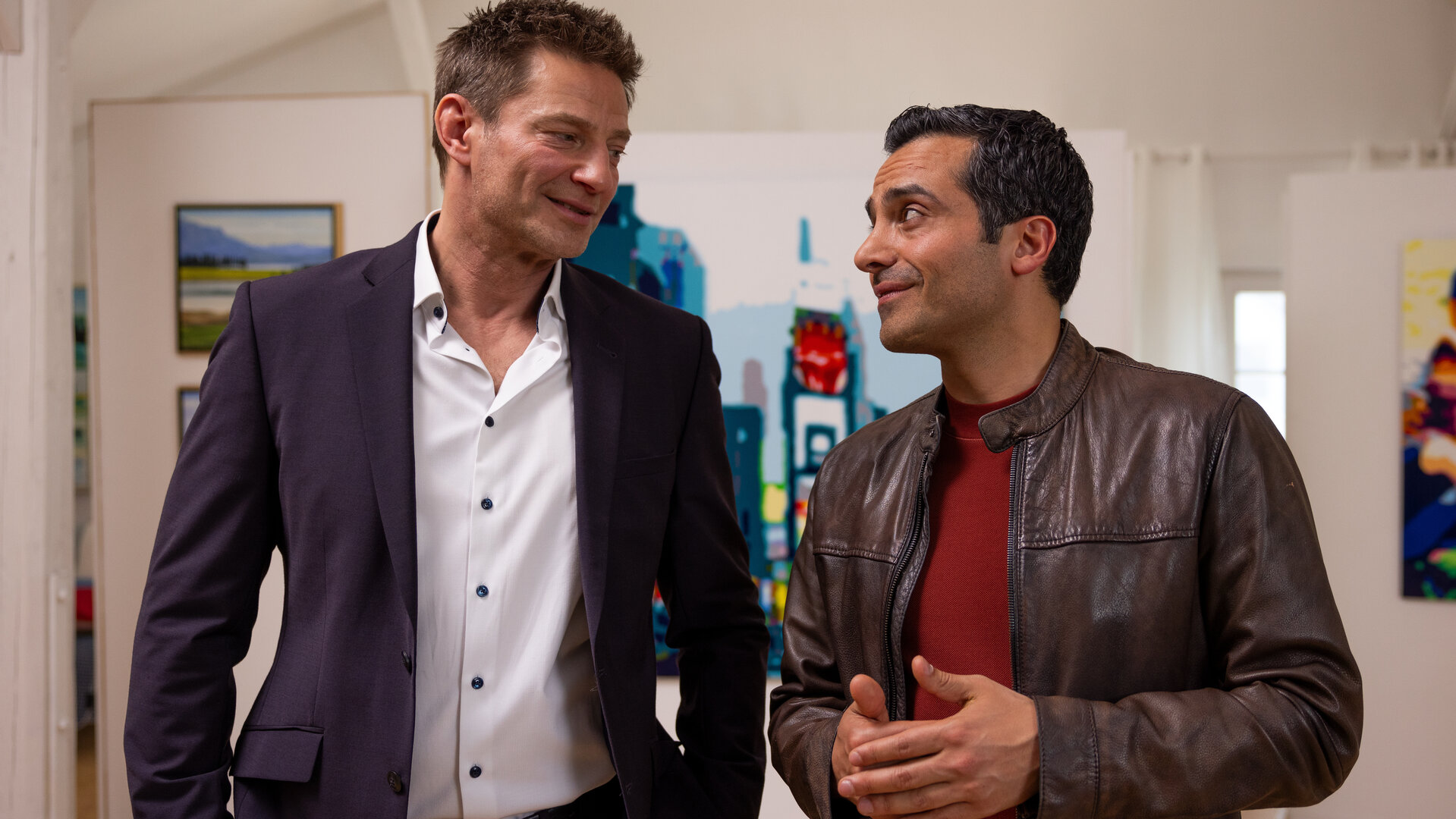 "Die Rosenheim-Cops - Original oder Fälschung": Die Kommissare Hansen (Igor Jeftić) und Kaya (Baran Hêvî) stehen lächelnd nebeneinander und sehen sich an. Im Hintergrund hängen diverse Gemälde an weißen Wänden.