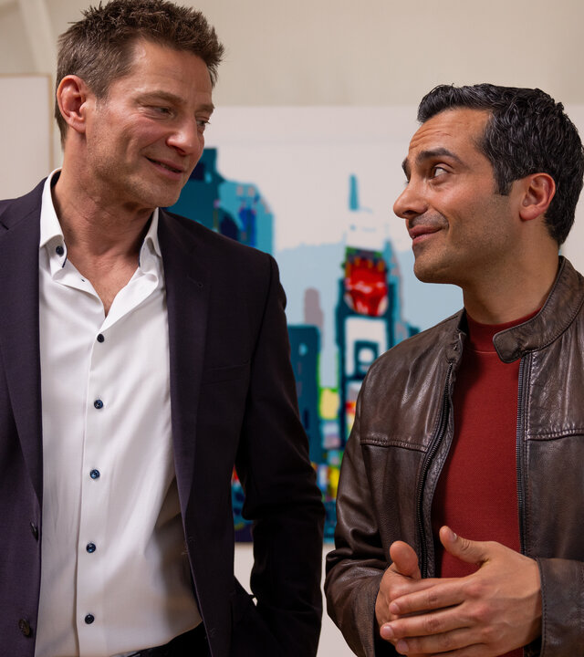 "Die Rosenheim-Cops - Original oder Fälschung": Die Kommissare Hansen (Igor Jeftić) und Kaya (Baran Hêvî) stehen lächelnd nebeneinander und sehen sich an. Im Hintergrund hängen diverse Gemälde an weißen Wänden.