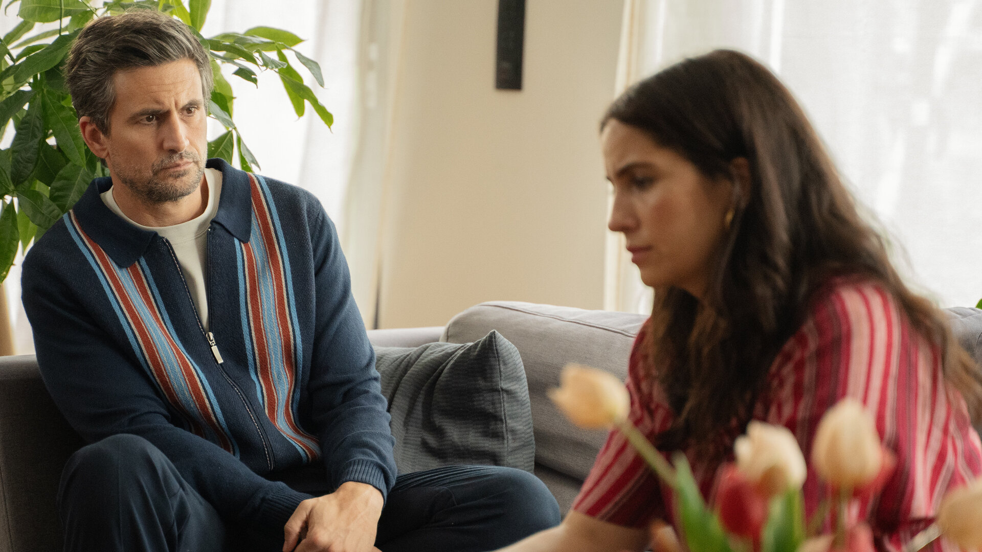 "The other gAIrl – Tischlein deck dich": Frank (Tom Beck) und Lisa (Chryssanthi Kavazi) sitzen nebeneinander auf dem Sofa. Frank blickt zu Lisa, die vor sich blickt.