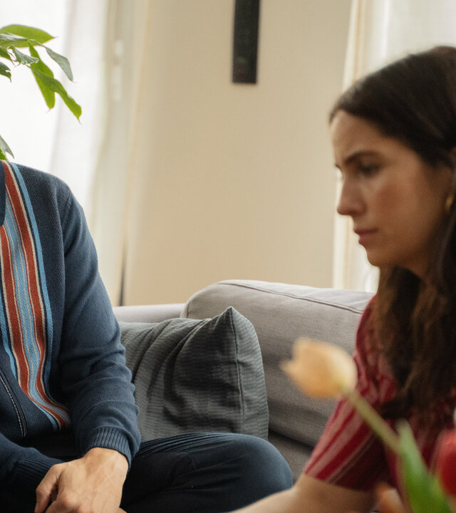 "The other gAIrl – Tischlein deck dich": Frank (Tom Beck) und Lisa (Chryssanthi Kavazi) sitzen nebeneinander auf dem Sofa. Frank blickt zu Lisa, die vor sich blickt.