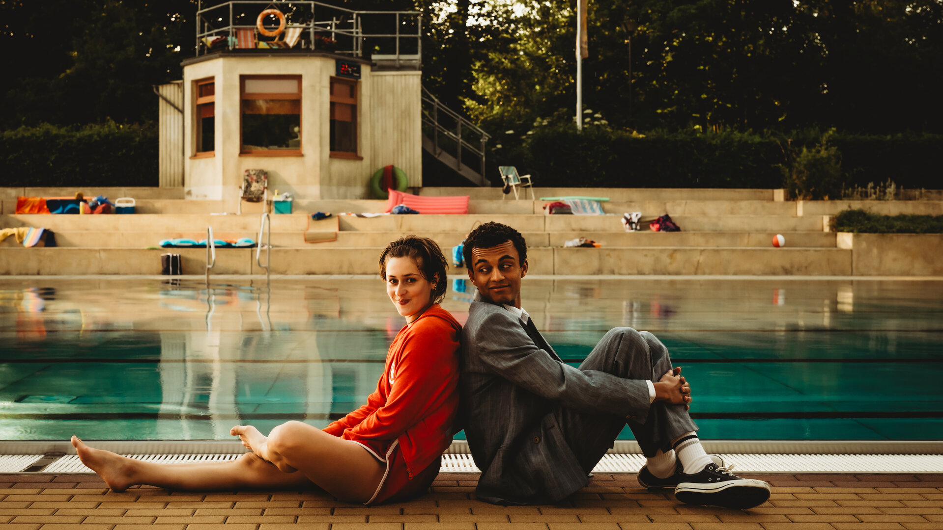 "Für immer Freibad": Mira (Anouk Elias) und Simon (Tyrell Otoo) sitzen Rücken an Rücken vor dem Becken des Freibads. Simon dreht seinen Kopf zu Mira und beide lächeln. Im Freibad sind keine weiteren Gäste.