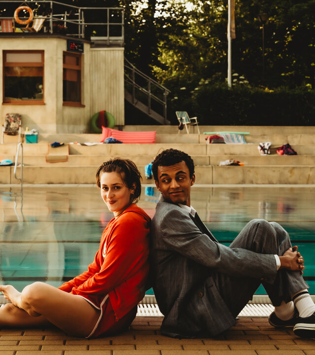 "Für immer Freibad": Mira (Anouk Elias) und Simon (Tyrell Otoo) sitzen Rücken an Rücken vor dem Becken des Freibads. Simon dreht seinen Kopf zu Mira und beide lächeln. Im Freibad sind keine weiteren Gäste.