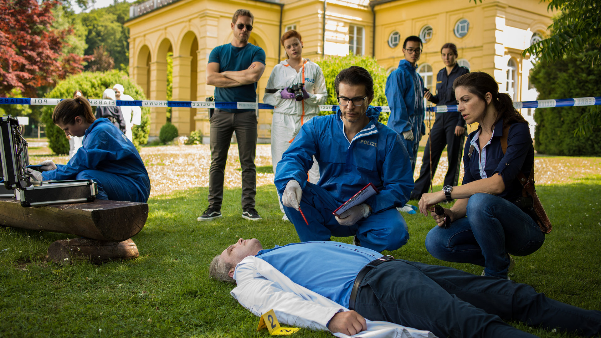 "SOKO München - Blindes Vertrauen": Am Tatort vor einer Villa liegt eine Leiche auf dem Rasen. Dr. Weissenböck (Florian Odendahl) und Katharina Hahn (Bianca Hein) sitzen in der Hocke neben ihr und schauen sie sich genauer an. Am Absperrband stehen Dominik Morgenstern (Joscha Kiefer) und Billie Curio (Sina Reiß), die eine Kamera in den Händen hält.