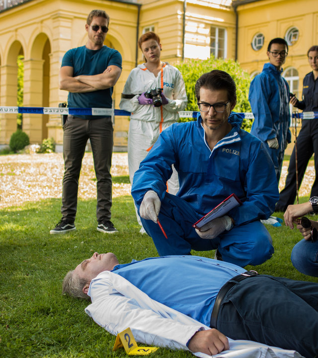 "SOKO München - Blindes Vertrauen": Am Tatort vor einer Villa liegt eine Leiche auf dem Rasen. Dr. Weissenböck (Florian Odendahl) und Katharina Hahn (Bianca Hein) sitzen in der Hocke neben ihr und schauen sie sich genauer an. Am Absperrband stehen Dominik Morgenstern (Joscha Kiefer) und Billie Curio (Sina Reiß), die eine Kamera in den Händen hält.