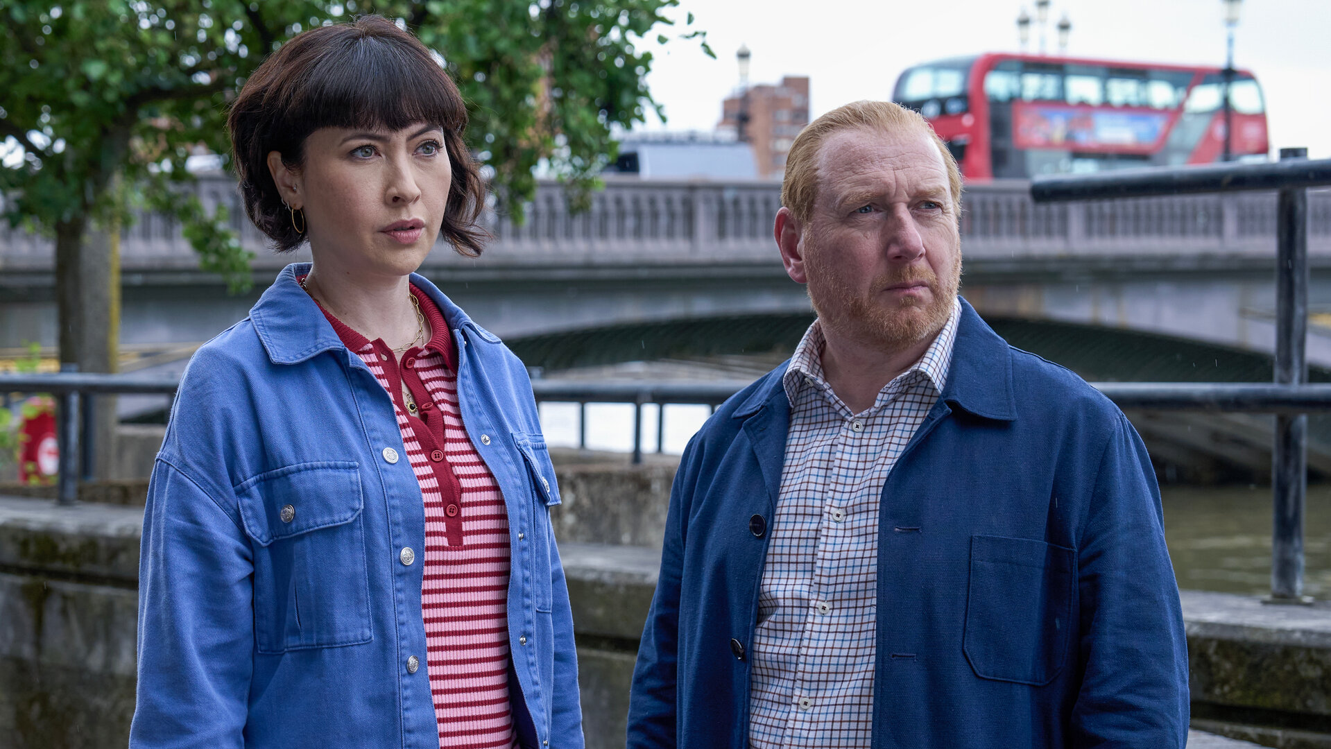 "The Chelsea Detective - Mythen und Legenden": DI Max Arnold (Adrian Scarborough) und DS Layla Walsh (Vanessa Emme) stehen nebeneinander vor einer Brücke über die Themse und schauen neugierig zur Seite. Ein roter Doppeldeckerbus fährt über die Brücke.