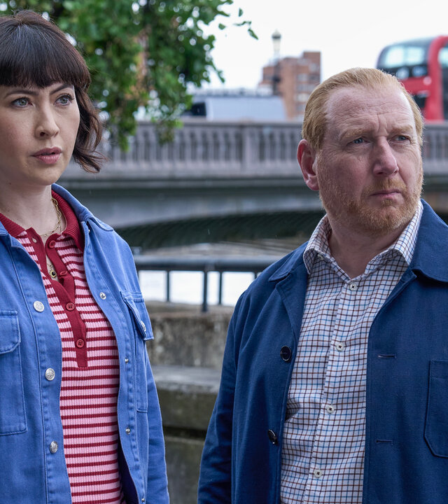 "The Chelsea Detective - Mythen und Legenden": DI Max Arnold (Adrian Scarborough) und DS Layla Walsh (Vanessa Emme) stehen nebeneinander vor einer Brücke über die Themse und schauen neugierig zur Seite. Ein roter Doppeldeckerbus fährt über die Brücke.