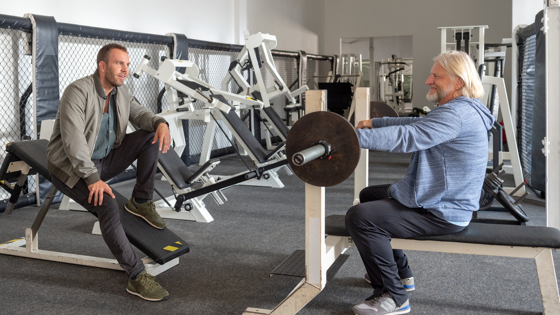 "SOKO Wismar – Sport ist Mord": In einem Fitness-Studio stehen verschiedene Trainingsgeräte in einer Reihe an der Wand. Studiobesitzer Bernd Peters (Frank Kessler) sitzt auf einer Hantelbank und stützt sich auf die Hantel. Kommissar Lars Pöhlmann (Dominic Boeer) sitzt ihm gegenüber, beide lachen sich an.