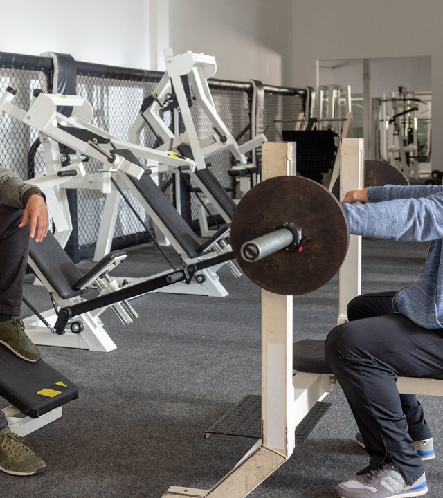 "SOKO Wismar – Sport ist Mord": In einem Fitness-Studio stehen verschiedene Trainingsgeräte in einer Reihe an der Wand. Studiobesitzer Bernd Peters (Frank Kessler) sitzt auf einer Hantelbank und stützt sich auf die Hantel. Kommissar Lars Pöhlmann (Dominic Boeer) sitzt ihm gegenüber, beide lachen sich an.