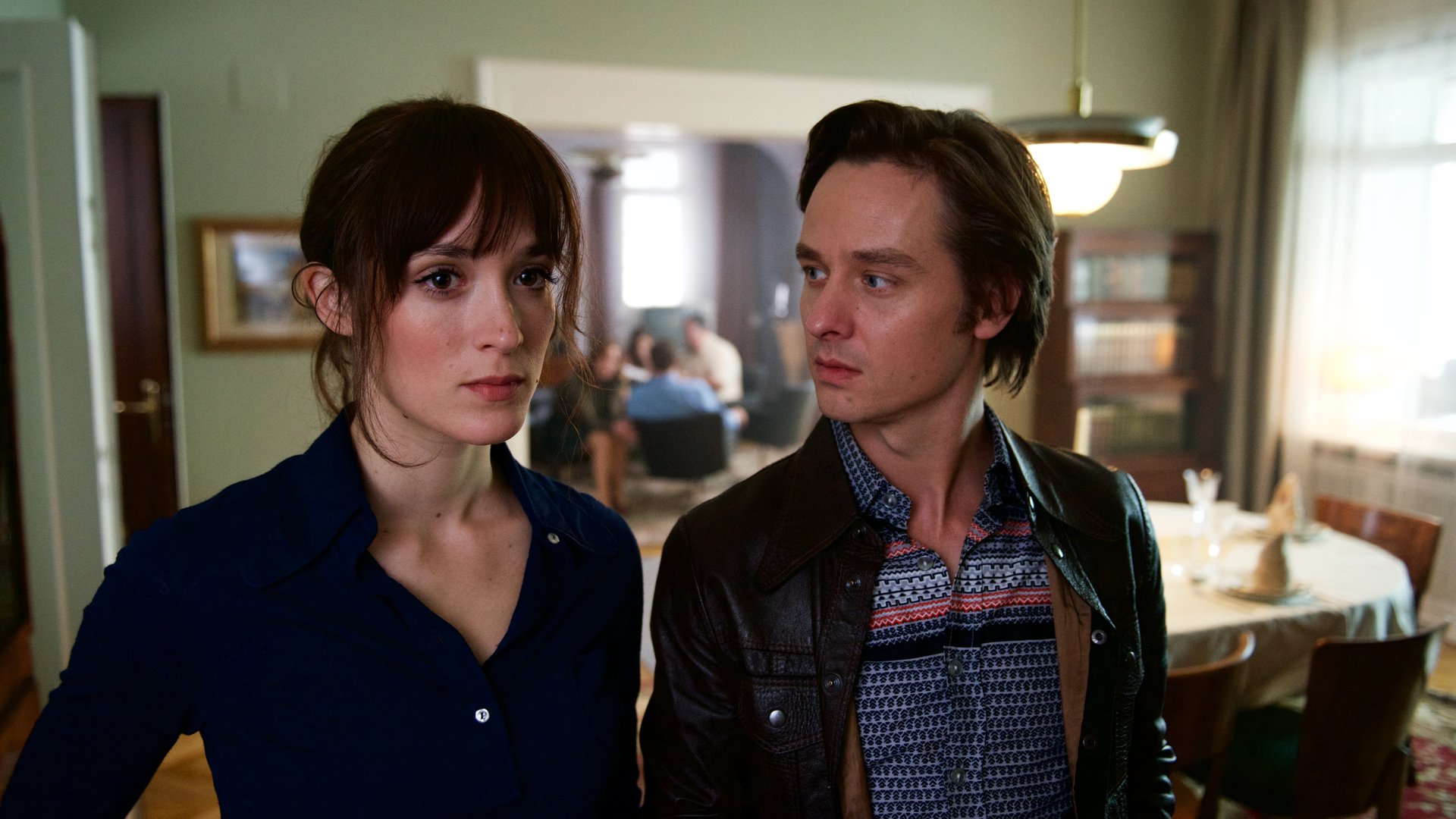 "Der gleiche Himmel (3)": Sabine Cutter (Friederike Becht) und Lars Weber (Tom Schilling) stehen Schulter an Schulter im Esszimmer der Familie Cutter. Lars schaut Sabine an. Im Hintergrund ist der Rest der Familie zu sehen.