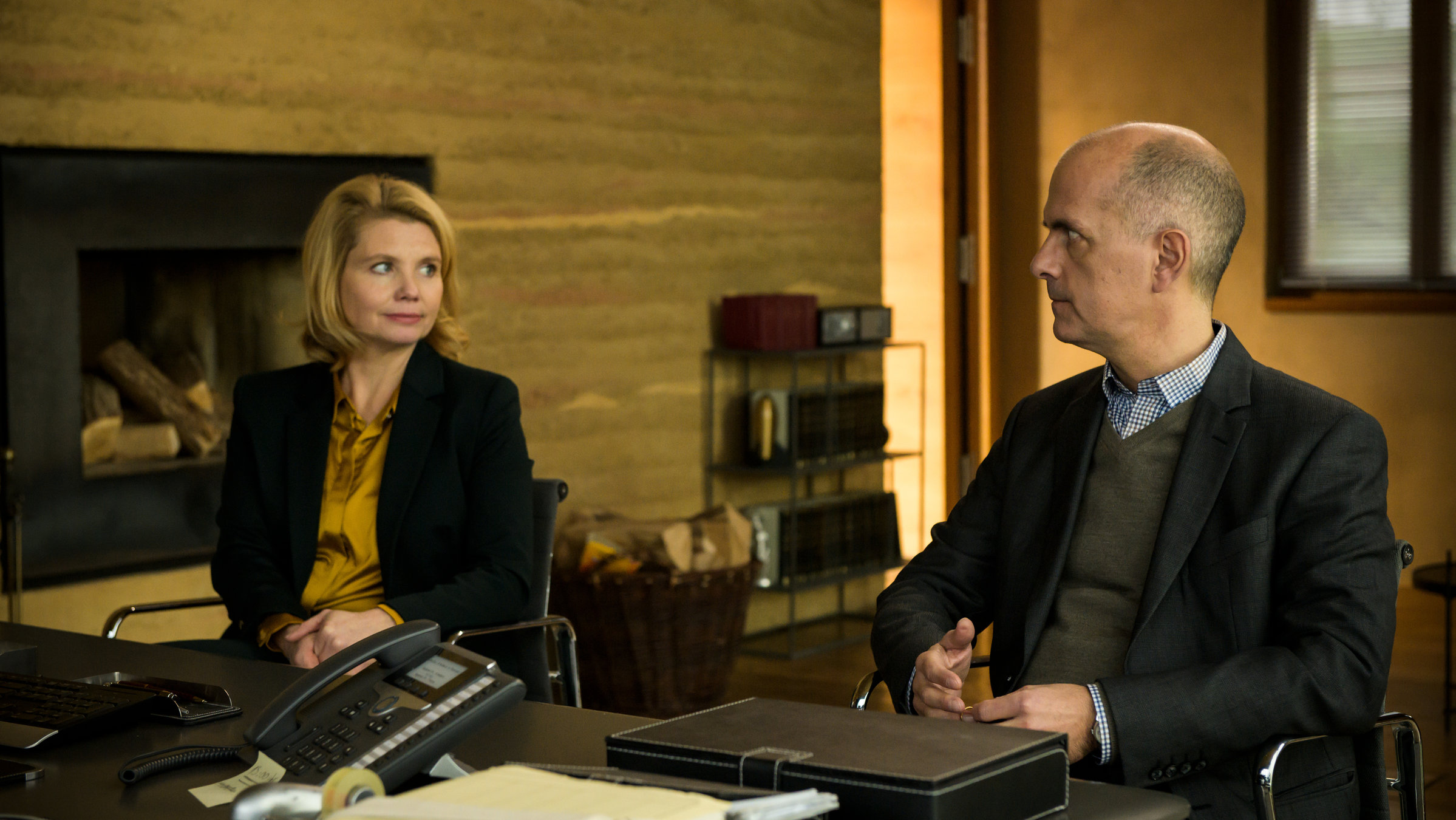 "Merz gegen Merz – Der Vertrag": Vor einem schwarzen Bürotisch sitzen Anne Merz (Annette Frier) und Erik Merz (Christoph Maria Herbst) nebeneinander und sehen sich gegenseitig aufmerksam an.