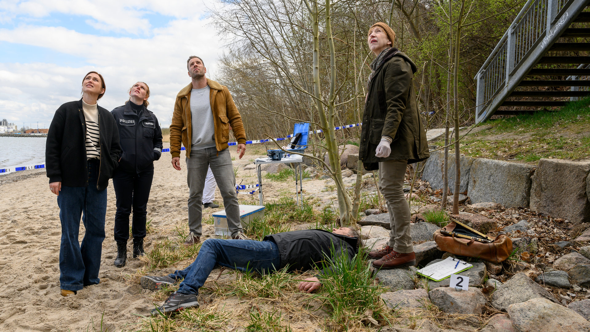 "SOKO Wismar – Lug und Trug": Am Fuße einer Seebrücke stehen Karoline Joost (Nike Fuhrmann), Paula Moorkamp (Stella Hinrichs), Lars Pöhlmann (Dominic Boeer) sowie Dr. Helene Sturbeck (Katharina Blaschke) um die am Boden liegende Leiche Holger Ahrens (Christian Wewerka) herum. Sie blicken nach oben auf die Seebrücke.