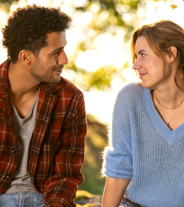 "Inga Lindström: Minnas Traum": Minna (Meriel Hinsching) und Lucas (Rojan Juan Barani) sitzen im Freien nebeneinander auf einer Holzbank und blicken sich lächelnd an. Im Hintergrund stehen Bäume an einem Gewässer.