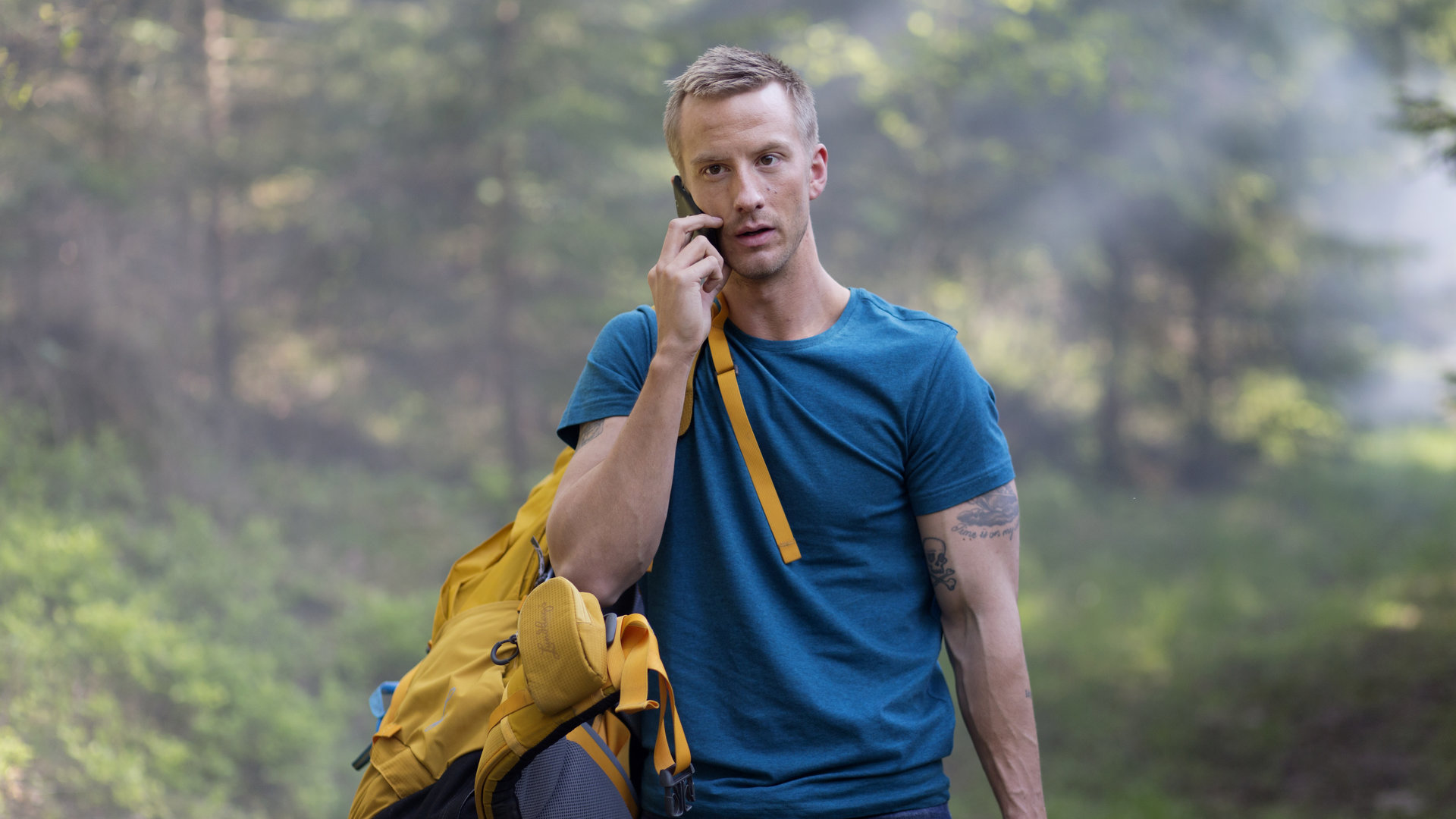 "Springflut - Vermisst": Ove Gardman (Filip Berg), mit einem blauen T-Shirt bekleidet und einem gelben Reiserucksack über der Schulter, läuft mit einem Handy telefonierend durch ein Waldstück.