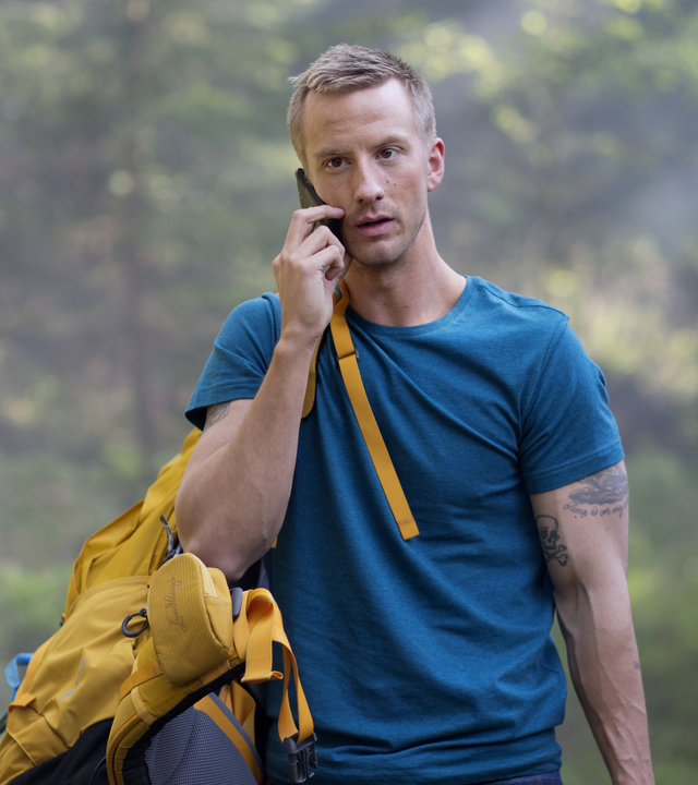 "Springflut - Vermisst": Ove Gardman (Filip Berg), mit einem blauen T-Shirt bekleidet und einem gelben Reiserucksack über der Schulter, läuft mit einem Handy telefonierend durch ein Waldstück.