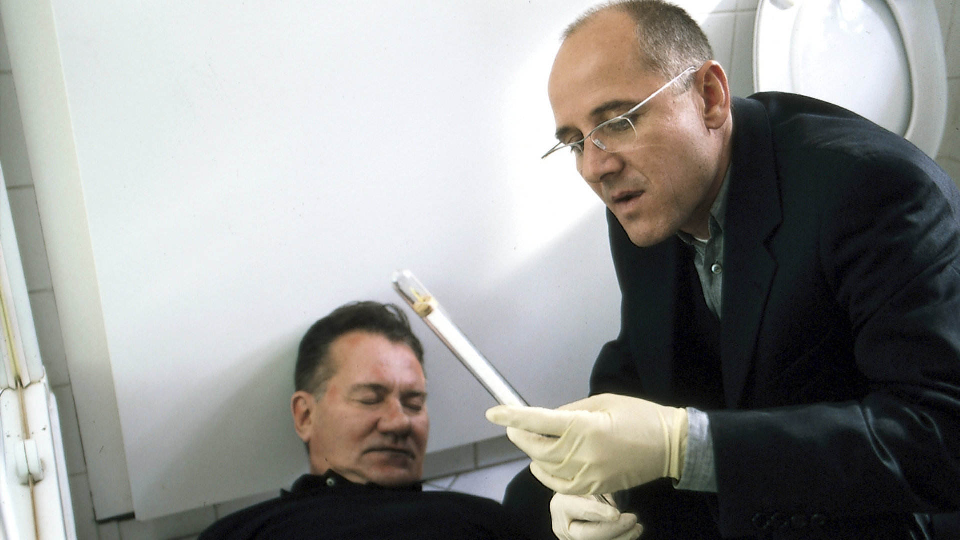 "Der letzte Zeuge - Die sich nach Liebe sehnen": Dr. Robert Kolmaar (Ulrich Mühe) begutachtet die Leiche von Jakob Fänger (Manfred Lehmann) am Tatort. Er schaut sich außerdem einen Gegenstand an.