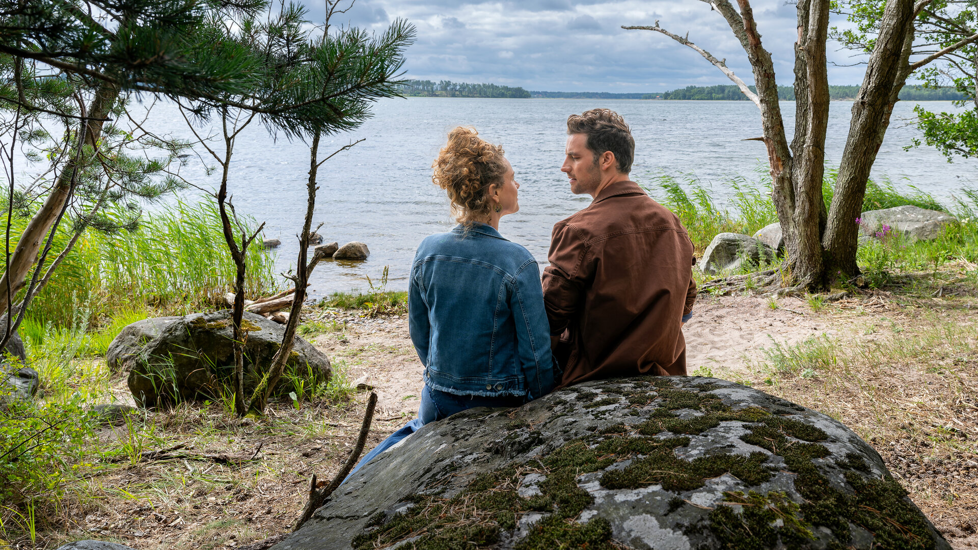 "Inga Lindström: Liebst du mich?": Jule (Klara Deutschmann) und Paul (Ferdinand Seebacher) sitzen mit dem Rücken zur Kamera auf einem runden Felsen an einem lauschigen Gewässerufer. Die beiden schauen sich an, vor ihnen stehen ein paar kleine Bäume und Schilf am Ufer.