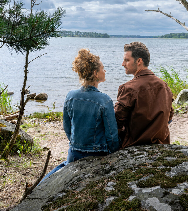 "Inga Lindström: Liebst du mich?": Jule (Klara Deutschmann) und Paul (Ferdinand Seebacher) sitzen mit dem Rücken zur Kamera auf einem runden Felsen an einem lauschigen Gewässerufer. Die beiden schauen sich an, vor ihnen stehen ein paar kleine Bäume und Schilf am Ufer.